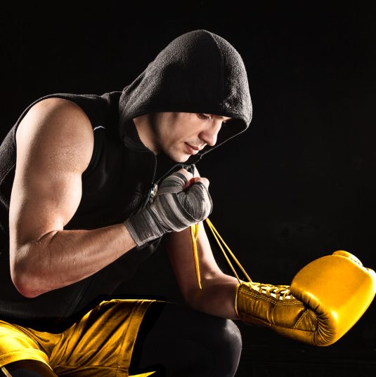 The young male athlete kickboxing sitting and lacing glove on a black  background
